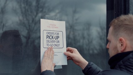 Caucasian male puts a Temporary closed due COVID-19 pandemic sign on a window. Coronavirus small business shutdown