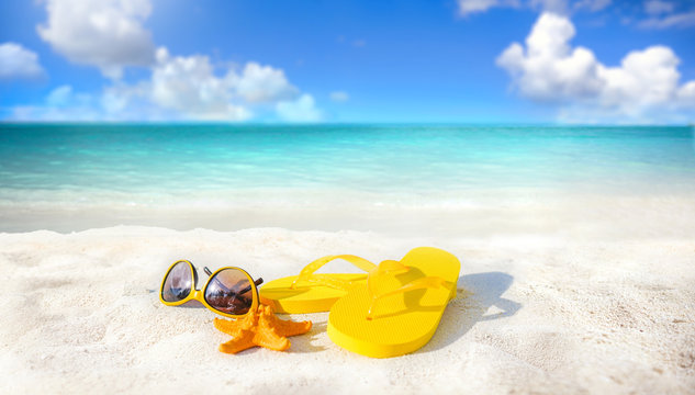 Concept of summer beach holiday. Beach accessories - sunglasses, starfish, yellow flip-flops on sandy tropical beach against blue sky with clouds on bright sunny day. Beautiful colorful image.