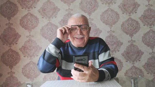 Old Man With Grey Mustache In Striped Jumper Looks At Modern Smartphone Screen Sitting At Table Against Outdated Wallpaper