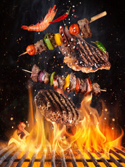 Tasty beef steaks and skewers flying above cast iron grate with fire flames. Freeze motion barbecue concept. - obrazy, fototapety, plakaty