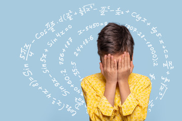 A young boy coveres his face with his palms experiences stress from an excess of information against complex mathematical expressions are written on a blue background.