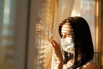 Woman with surgical mask looking out of home window, concept of quarantine during viral pandemic...