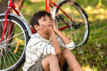 Obraz premium A young boy sits on the grass in the garden and dreams of something. A red bike is standing by a tree, a book is lying on the grass