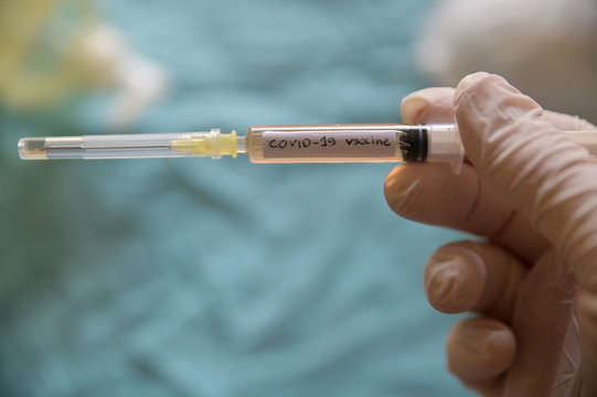 Close up shot of a syringe with the word coronavirus vaccine.He is holding her hand with a white latex glove.In the blurred background a white mask and the spout of the sanitizing gel.Conceptual image
