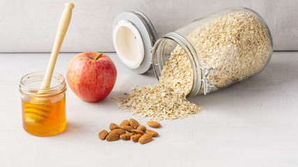 Transparent oatmeal jar with oats spread out on grey background. There is an apple, some almonds and honey pot near it. Grey background. 