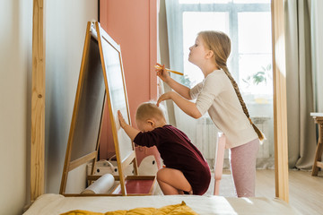 The sisters play together in their large, sunny and bright room. They do not leave the house in self-isolation mode in an epidemic. Girls draw with felt-tip pens on a blackboard.  Kids play and smile.