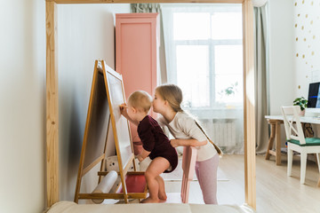 Kid's play together in their large, sunny and bright room. They do not leave the house in self-isolation mode in an epidemic. Girls draw with felt-tip pens on a blackboard.  Kids play and smile.