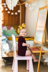 Little girl draws with felt-tip pen on a blackboard in a bright and stylish children's room. Baby sitting on a chair and smiling