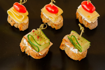 One bite sandwiches with ham, cucumber, cheese, and tomatoes on a black background. 