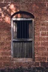 Arched dark brown wooden window closed off by metal bars sits in the middle of a exposed brick wall.  