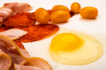 A broken chicken egg without a shell, slices of bacon and sausage and tomatoes on a white background. Close up.