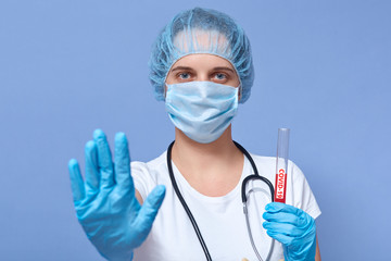 Indoor shot of young beautiful girl wearing rubber gloves, medical mask and disposable hat, holding laboratory blood sample of coronavirus disease, showing stop sign with hand, has serious expression.