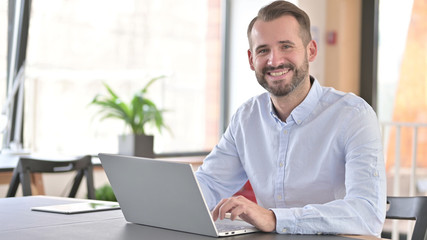 Young Man with Laptop Smiling at Camera in Office