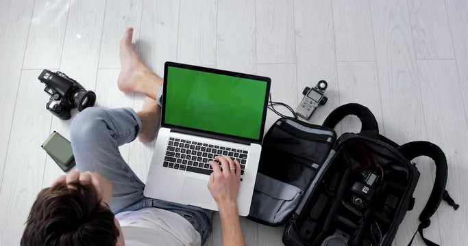 Top View of a Young Hipster Photographer Editing Photos on MacBook Pro Green Screen. Backpack, Medium format film and DSLR Camera, Lens, External Hard Drives and Light Meter. Freelancer Working Home