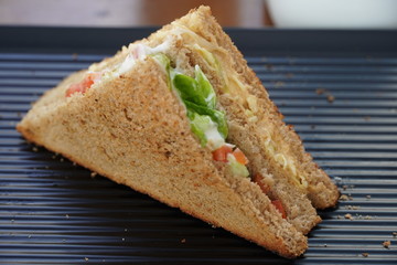 A single Tuna sandwich on black serving plate. One Club Sandwich with chicken, ham, cheese, tomatoes, cucumber, bacon, lettuce, herbs and toasted bread. Front view with blurred background.