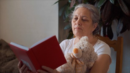 Quarantine senior woman reading book rest with teddy bear toy. Home isolation. 