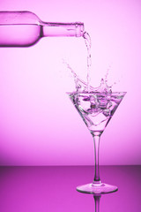 Pink bottle spilling liquid into a cocktail glass and making splash. Pink background, alcoholic drinks and cocktails concept.