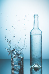 Bottle of ice water with drops, and glass of water with ice falling with splash. Water splash and Refreshing Drinks Concept.