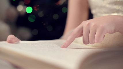 Close-up soft focus the hand of a little girl leads along the lines of a book while reading a New Year's fairy tale amid the lights of a Christmas tree. The concept of a fabulous mood