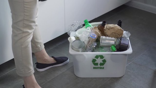 American woman is throwing trash and showing recycling bin in home interior. Spbd Young housewife female is putting used garbage in container with green ecological sign for recycle while standing in