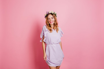 Slim girl in beautiful purple dress laughing on bright background. Studio shot of cute blonde woman with fresh flowers in hair enjoying photoshoot.