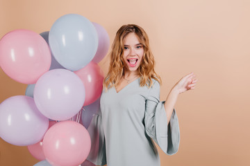 Fototapeta na wymiar Charming fair-haired birthday girl posing with pleasure on light background. Indoor photo of spectacular woman dancing with balloons at event.