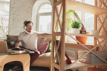 Young man studying at home during online courses for laborer, journalist, developer. Getting profession while isolated, quarantine against coronavirus spreading. Using laptop, smartphone, headphones.