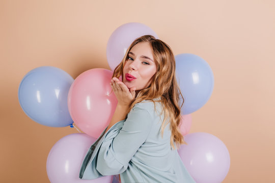 Lovely birthday girl with wavy hair posing on light background. Studio shot of positive lady sending air kiss and holding blue balloons.