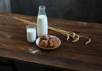 On a wooden table is a bottle and a glass of milk, cookies with chocolate on a plate, spikelets of wheat