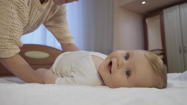 Careful mother changes baby diaper at home. Cute toddler with blue eyes lies on a diaper in bedroom, mom and toddler concept of a happy childhood. Low-angle shot in close-up