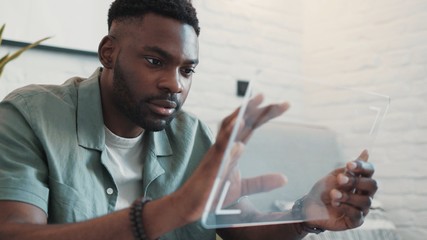 Young african-american man sits on sofa uses futuristic transparent tablet works visual effects home modern screen tech abstract application communication innovation close up