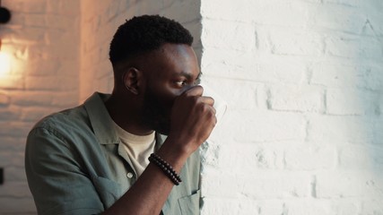 Close up view of attractive african-american man stays near the window wathcing through the window on great view drinking coffee morning modern room relaxen enjoys day