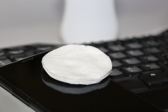 Round pledget / swab on smartphone screen and compuer keyboard close up, in background white pharmaceutical bottle with cleaning liquid