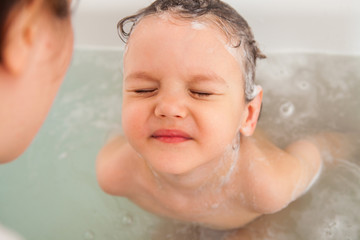 Mom washes her baby's head in a bath with water. Flushing shower shampoo foam. Shampooing without tears.