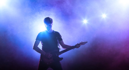 Young male singer performing on a stage and playing the guitar. Smoky blue background - obrazy, fototapety, plakaty