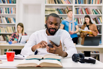 Young handsome African happy man, student, with beard and in white shirt, sitting at the table with books in library reading room and using tablet pc, looking for information for study