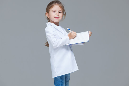 Adorable Child Girl Uniformed As Doctor Is Writing Medical Record Isolated On Grey Background