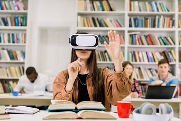 Young pretty girl, student, library user, using VR simulator while doing research in library. Woman wearing virtual reality glasses, sitting at desk with many different books and preparing for exam