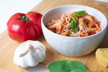Classic italian penne pasta arabiata with its ingredients in the background: fresh tomatoes, basil, garlic and parmesan cheese