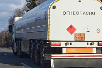 Close up white semi truck fuel tanker with 33 1203 dangerous class sign and inscription in Russian INFLAMMABLE moving on asphalt highway on a spring day, side rear view - ADR hazardous cargo