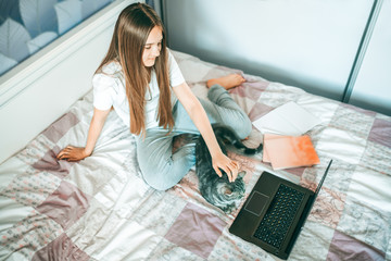 A schoolgirl with her cat pet holding a video conference with a tutor on a laptop at home during quarantine, sitting on the bed. The concept of distance learning.