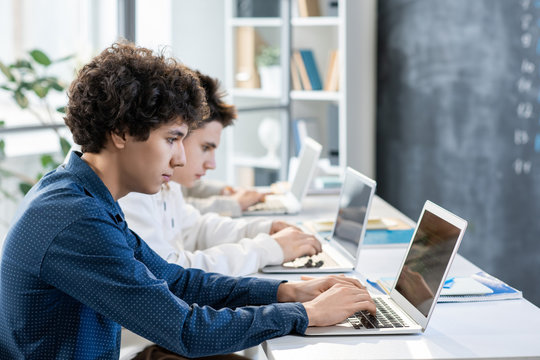 Side view of two guys sitting by desk in row in front of laptops