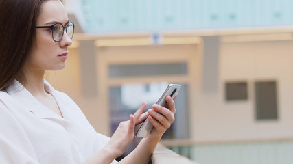 young woman in a white shirt and glasses uses smartphone to search for information on the Internet.