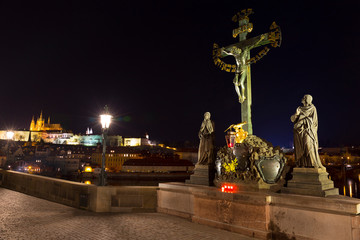 Fototapeta na wymiar Baroque statue of Jesus Christ on the Prague Charles Bridge with the gothic Castle in the Night without People at the time of Coronavirus, Czech republic