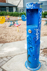 A modern designed blue drinking fountain with a park in the background, tag-graffiti and selective focus