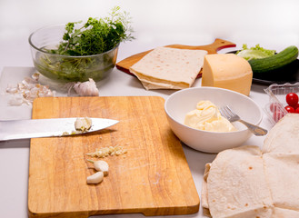 Cutting board with a knife, a bowl with greens, a black plate with a lettuce, a bowl with sauce, cherry tomatoes, a piece of cheese, sliced ​​garlic on a white plate, close-up