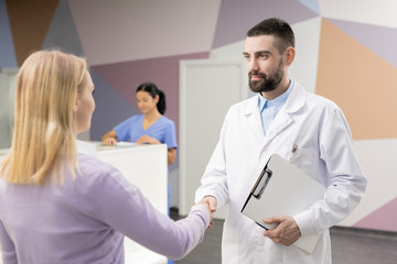 Fototapeta premium Young confident doctor in whitecoat handshaking with one of patients