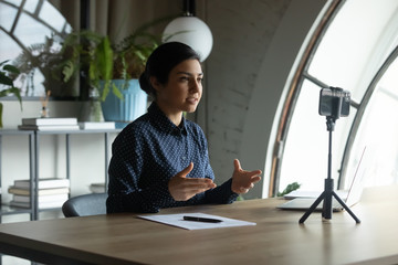 Focused young indian ethnicity businesswoman sitting at table in front of cellphone on stabilizer, recording educational video for personal vlog, sharing knowledge with followers online in office.