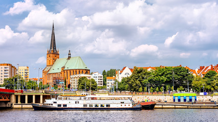 Ships anchored on Odra River pier. People relaxing on Piastowski Boulevard. Cathedral Basilica of St James the Apostle in background, Szczecin, Poland - obrazy, fototapety, plakaty