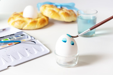 Brush is painting blue dots with watercolor on an Easter egg for the holiday decoration, light grey background
